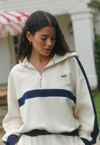 Krémová mikina na zips s tmavomodrým vodorovným pruhom, mäkkou textúrou a voľným strihom. Obsahuje logo „self.“ a kontrastné tmavomodré manžety a lem.