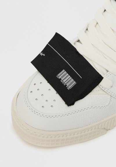 Białe sneakersy z fakturowaną skórzaną cholewką, perforowanym noskiem i czarną tkaniną metki z kodem kreskowym. Sznurówki są w jasnym odcieniu beżu.