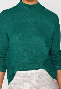 Turquoise gebreide trui met een hoge kraag, lange mouwen en geribbelde boorden. Zachte textuur en een ontspannen pasvorm, geaccentueerd door zijsplitten aan de zoom.