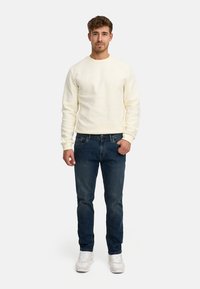 Jonge man staat, draagt een crèmekleurige trui met structuur, donkerblauwe spijkerbroek, witte sneakers, met één hand in zijn zak tegen een effen achtergrond.