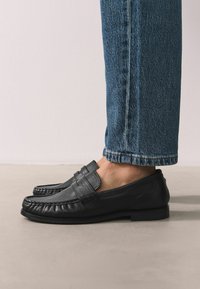 Black leather loafers with stitched detailing and a flat sole, paired with cuffed blue denim jeans. Shoes feature a slip-on design.