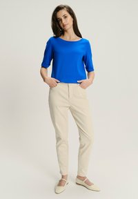 Kobieta stoi z rękami w kieszeniach, ubrana w niebieską bluzkę z krótkim rękawem, beżowe spodnie i jasnobeżowe klapki na płaskim obcasie na jednolitym tle.