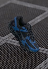 Black and blue Nike sports shoe with reflective laces on a metal grid surface.