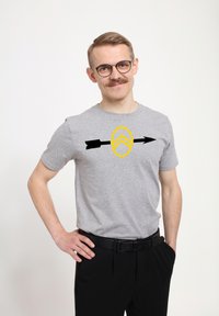 Šedé bavlněné tričko s žlutým štítem, na kterém je grafika chevronu a černé šipky. V kombinaci s černými kalhotami a opaskem.