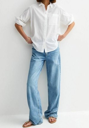 Personne portant une chemise blanche à boutons avec les manches retroussées, un jean ample bleu clair et des sandales beige, debout les mains sur les hanches.