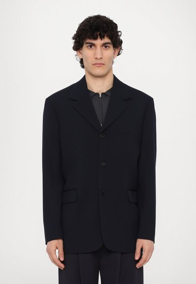 Blazer azul marino oscuro con un diseño estructurado, dos bolsillos frontales, solapas con muesca y tres botones. Lleva puesto un top gris con cremallera.