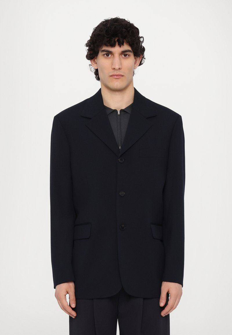 Blazer de un băiețel închis, cu un design structurat, două buzunare frontale, rever cu un unghi și trei butoane. Purta un top gri cu fermoar.