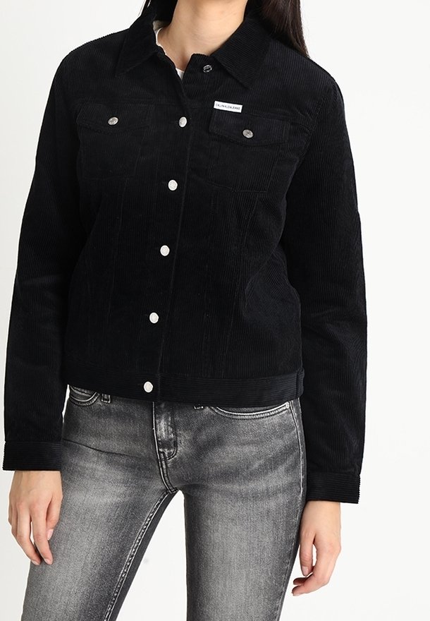 Černá kordurová bunda s špičatým límcem, dvěma náprsními kapsami, stříbrnými doplňky a zkráceným střihem, kombinovaná se šedými džíny.