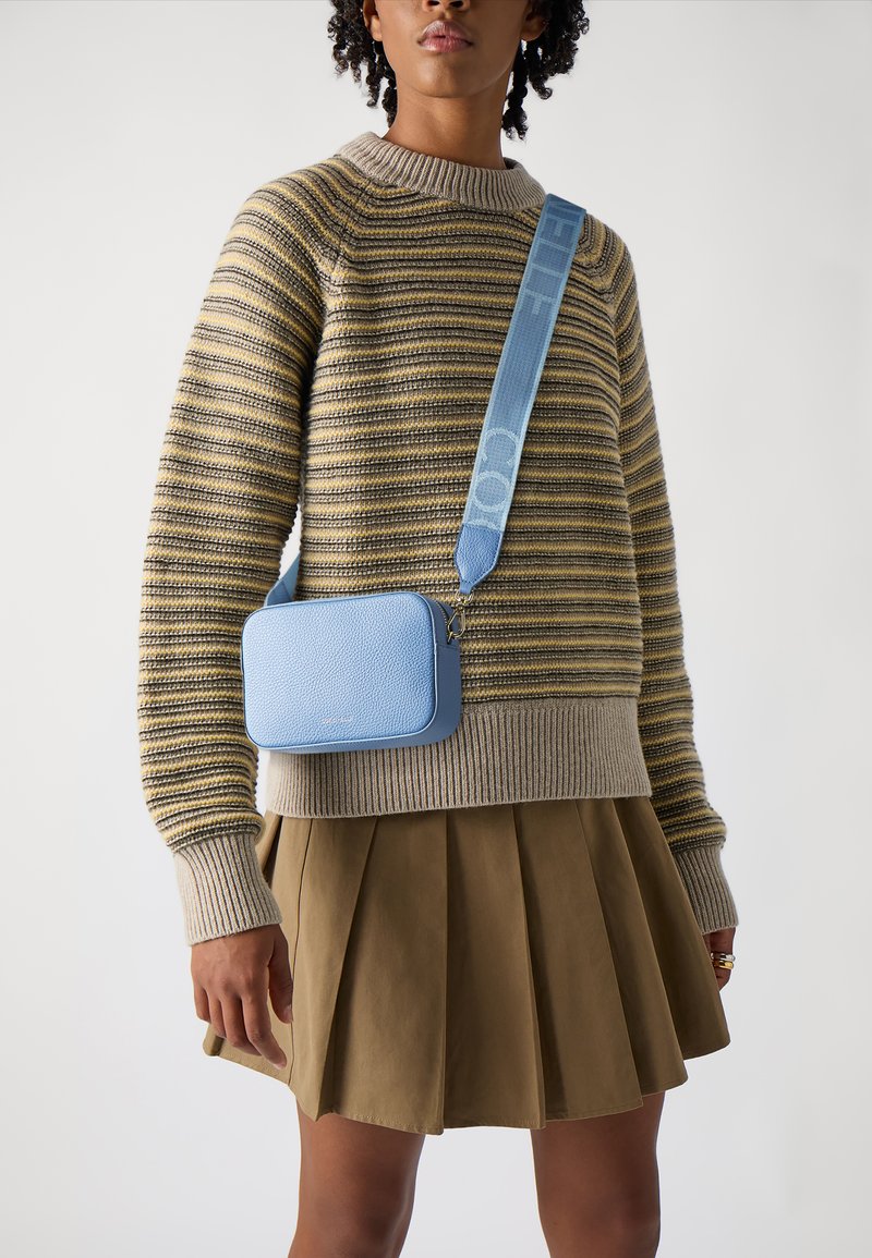 Světle modrá kožená crossbody taška s texturovaným povrchem, obdélníkového tvaru, a širokým popruhem s vzorem, nošena přes pruhovaný svetr a sukni.