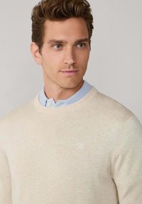 Pull en tricot beige avec un col rond et une finition texturée, associé à une chemise à col bleu clair en dessous et un petit détail de logo blanc.