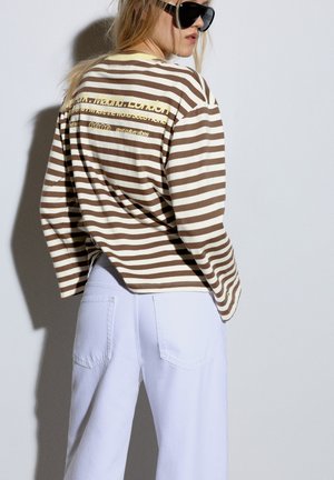 Blonde Frau mit schwarzen Sonnenbrillen, einem braun-weiß gestreiften Langarmshirt mit Text und hoch taillierten weißen Hosen, seitlich gesehen.