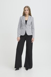 Vrouw staand, gekleed in een grijs nauwsluitend colbert, zwarte broek met wijde pijpen en zwarte pumps met spitse neus, tegen een effen lichte achtergrond.