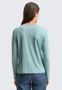 Woman with long brown hair wearing a light blue long-sleeve top and blue jeans, shown from the back against a plain background.