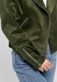 Giacca da motociclista in suede verde oliva con zip argentate su maniche e tasche frontali, dal design aderente e superficie texture.