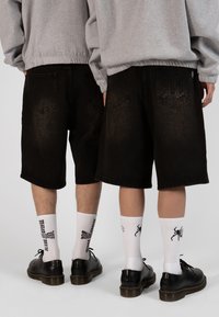 Shorts en denim noir avec un effet délavé, dotés de deux poches arrière et d'un détail de logo. Portés avec des chaussettes blanches et des chaussures noires.