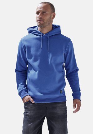 Blue hoodie made of soft fabric, featuring a drawstring hood, ribbed cuffs, and a small logo patch near the hem. Worn with dark jeans.