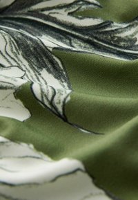 Žalia audinys su gėlių raštu juodomis kontūromis. Tekstūra yra lygi ir šiek tiek blizgi, su švelniais, natūraliais klostės.
