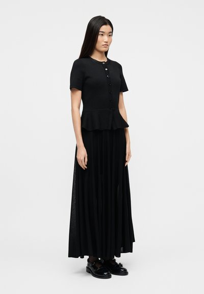 Γυναίκα στέκεται φορώντας ένα μαύρο πουκάμισο με κοντά μανίκια και κουμπιά, με πεπλούμιο τελείωμα, και μια μακριά μαύρη πλισέ φούστα με μαύρα παπούτσια.