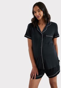 Czarny zestaw piżamowy z miękkiego materiału, składający się z krótkiego topu z guzikiem i krótkim rękawem, wykończonego białym lamowaniem oraz kieszonką na piersi, w połączeniu z pasującymi szortami.