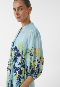 Hellblaues Kleid mit floralen Druck in Blau, Gelb und Grün. Es hat einen Mandarin-Kragen und Puffärmel und besteht aus einem leichten Stoff.