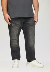 Jean en denim noir avec un design usé, présentant des patches décolorés et une coupe décontractée, associés à des baskets blanches et un t-shirt gris.