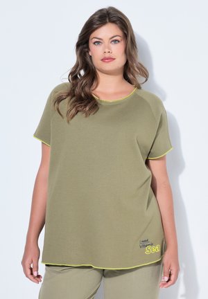 Donna con capelli castani mossi che indossa una maglietta a maniche corte verde oliva e pantaloni, con la scritta "Ho bisogno di vitamina mare" sull'orlo della maglietta.