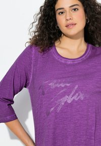 Fioletowa koszula z długim rękawem i okrągłym dekoltem, ozdobiona błyszczącym napisem w stylu pisma odręcznego. Tkanina ma fakturę z efektem melanżu.