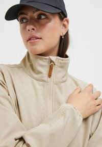 Beige high-collared jacket with smooth texture, zip closure, and logo detail on sleeve. Model wears a black cap and subtle earrings.