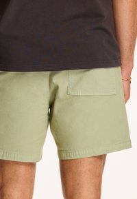 Shorts i lys grøn bomuld med en baglomme og en grov overflade. Sammen med en mørk t-shirt med korte ærmer.