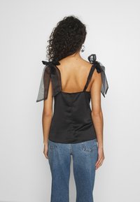 Svart satin topp med genomskinliga svarta band som är knutna till rosetter. Har en figurnära passform och dragkedja i ryggen, kombinerad med blå jeans.