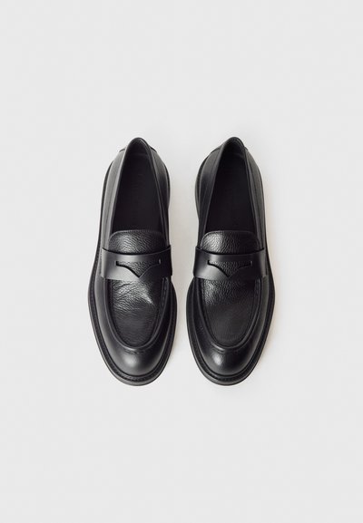Sapatos mocassins de couro preto com acabamento liso, biqueira arredondada e uma tira decorativa no peito do pé. Sola plana com detalhes sutis de costura.