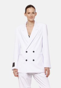 Kobieta z gładko zaczesanymi włosami nosi oversize'ow�ą, podwójnie zapinaną białą marynarkę z czarnymi guzikami oraz dopasowane białe spodnie na jednolitym tle.
