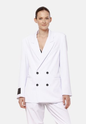 Kobieta z gładko zaczesanymi włosami nosi oversize'ową, podwójnie zapinaną białą marynarkę z czarnymi guzikami oraz dopasowane białe spodnie na jednolitym tle.