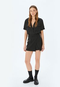 Camisa negra de botones con mangas cortas combinada con una falda envolvente negra que tiene un bolsillo lateral y detalles de botones dorados. Calcetines negros y mocasines.