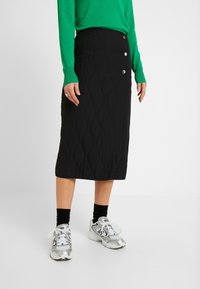 Jupe midi matelassée noire avec un motif texturé et trois accents de boutons argentés, portée avec un haut vert et des chaussures de sport argentées.