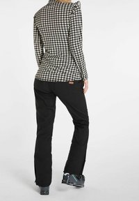 Persona che indossa pantaloni da sci neri, un top a scacchi bianco e nero e scarponi da sci grigi, in piedi con la schiena rivolta in avanti su uno sfondo bianco.