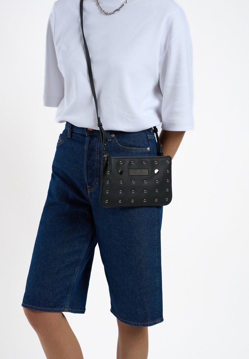Zwarte leren crossbody tas met zilveren studs, rechthoekige vorm, gladde textuur en verstelbare strap, gedragen met denim shorts en een witte top.