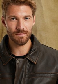 Brun läderjacka med krage, synlig hårdvara vid nacken och strukturerad yta med kontrasterande sömmar. Närbild av jackans detaljer.