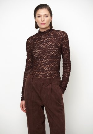 Vrouw met kort haar draagt een donkerbruine lange mouwen top met bloemenkant en een hooggetailleerde bruine broek tegen een witte achtergrond.