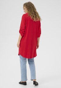 Röd långärmad blus med uppvikta ärmar, rundade fållar och mjuk textur. Matchad med ljusblå jeans och svarta platta skor.