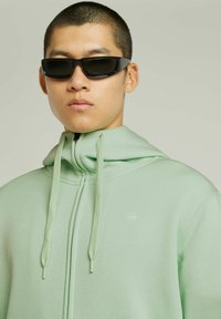 Haut de sweat à capuche zippé vert clair avec un col montant, des cordons de serrage et un petit logo. Modèle portant des lunettes de soleil noires élégantes. Texture de tissu lisse.