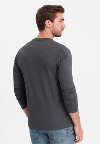 Mies, jolla on lyhyet ruskeat hiukset, pukeutunut tummanharmaaseen pitkähihaiseen paitaan ja sinisiin farkkuihin, seisoo ja katsoo oikealle valkoista taustaa vasten.