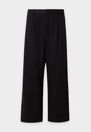 Høj mand iført en mørkeblå åben skjorte over en hvid t-shirt, brede sorte bukser og sorte sko, stående mod en ensfarvet grå baggrund.