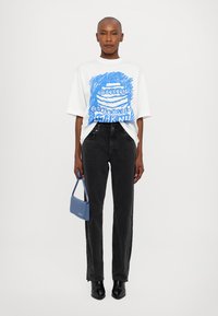 T-shirt bianca con design grafico blu, abbinata a jeans neri a gamba dritta, stivali neri alla caviglia e piccola borsa a tracolla azzurro chiaro.