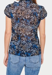 Seorang wanita mengenakan blus hitam lengan pendek semi-transparan dengan motif bunga biru, krem, dan putih serta celana jeans denim biru muda, dilihat dari belakang.