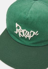 Gorra de pana verde con visera plana, que presenta texto bordado en blanco en la parte frontal. La textura es acanalada y suave.