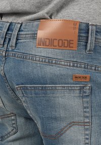 Plave traperice s smešnim kožnim oznakom na kojoj piše "INDICODE." Sadrže vidljive šavove i klasičan dizajn džepova.