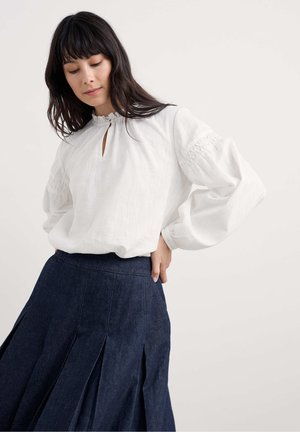 Vrouw in een witte blouse met lange mouwen en een smockmouw, gekleed in een donkere geplooide denimrok, staande tegen een effen achtergrond.