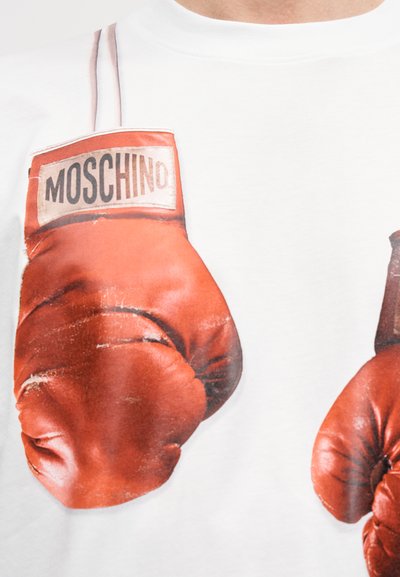 Λευκό μπλουζάκι με μεγάλο γραφικό κόκκινων γαντιών πυγμαχίας, με ορατή ετικέτα της μάρκας "Moschino" και φθαρμένη υφή στα γάντια.