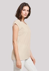 Beige Kurzarm-V-Ausschnitt-Oberteil aus weichem Stoff, lockere Passform, mit glatter Textur und schlichtem Design. Getragen mit weißen Hosen.